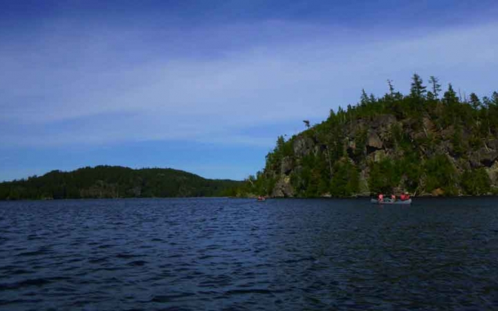 a canoe is paddled on a calm blue lake along side a tree-lined shore 